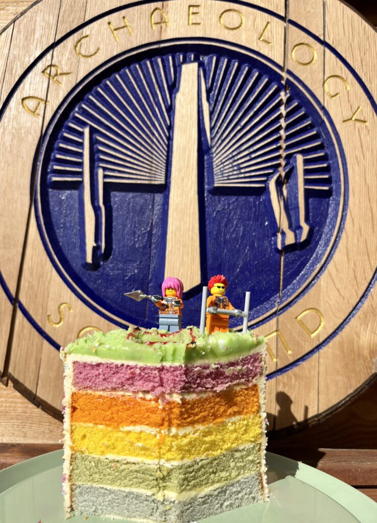 A large slice of rainbow layered cake on a green plate. On the green icing on top stand two Lego figurines holding tools next to a hole. In the background is a big wooden round sign with a carved design painted blue and gold lettering.