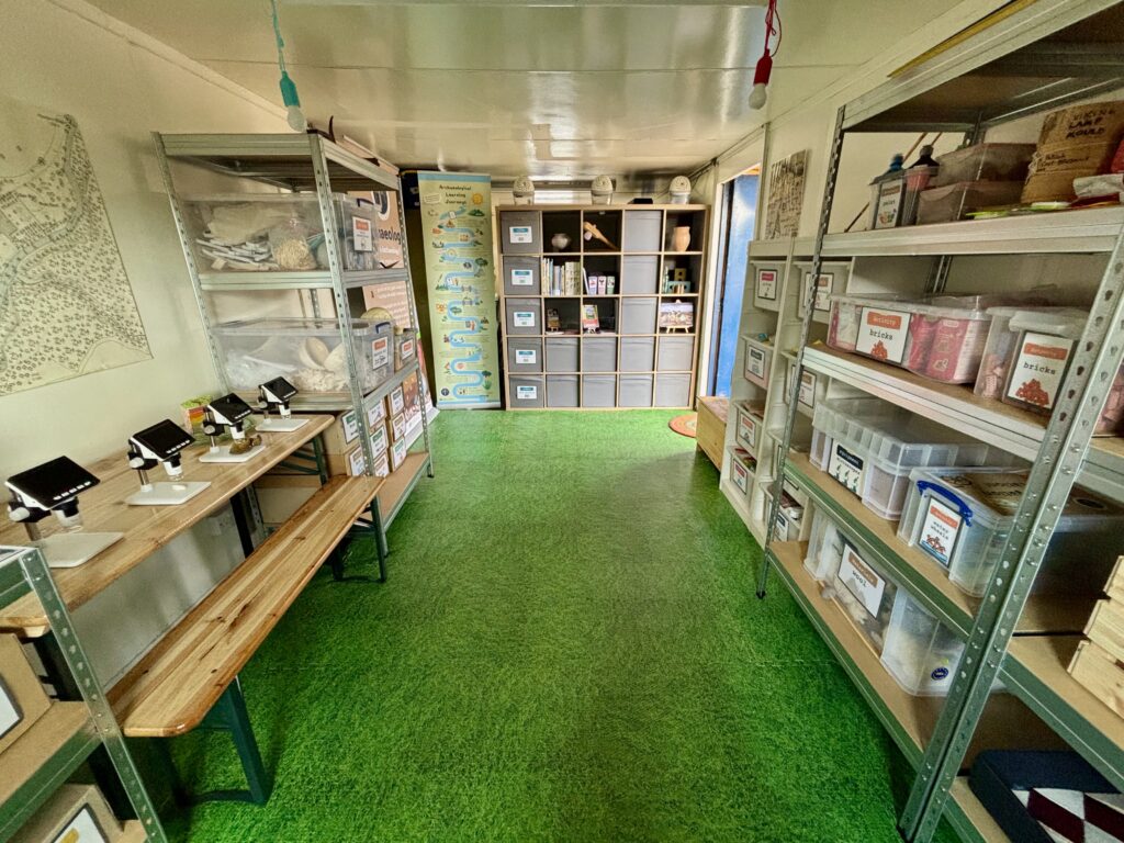 The inside of a building with green grass effect flooring and a shiny white ceiling with two bulbs hanging down. There are shelving units with labelled boxes, as well as a table and bench with microscopes, There is a large banner in the background, and an old map on the left wall. The blue door at the back right is open and light is shining in.