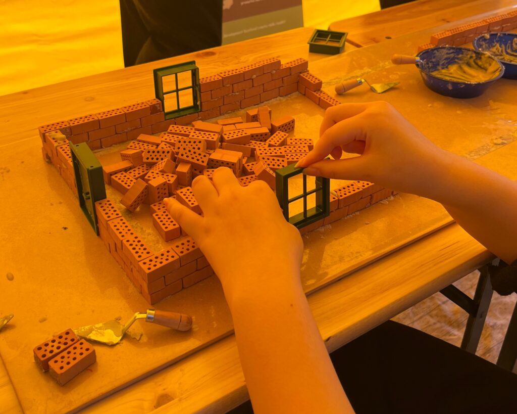 Two hands building the foundations of a small building made from miniature bricks, with windows and a door. The building is on a wooden table which has small trowels and bowls of mortar. There is a yellow tint to the photo from the gazebo in the background.