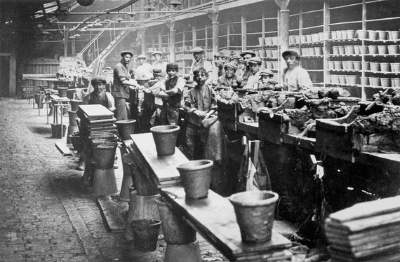 A black and white photo shows three flowerpots on planks. Behind the pots there are people standing facing the camera. They are boys and men and lots are wearing hats. Behind them are shelves with pots on, and lighting above their heads.