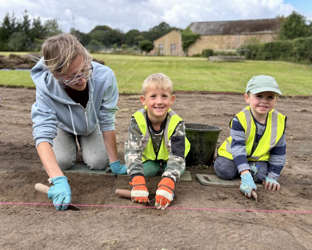 An adult and two children in hi-vis yellow vests are kneeling on the ground. They are wearing orange and blue gloves and holding trowels and smiling.