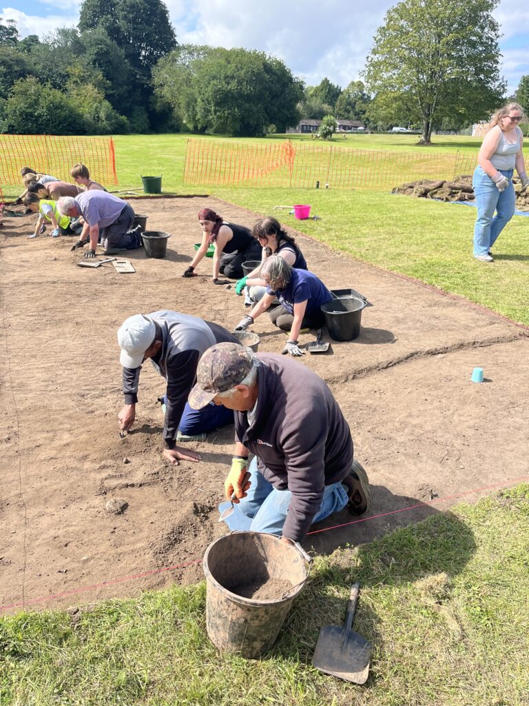 People kneeling in a row on a sunny day. There is a man with a cap and a bucket next to him.