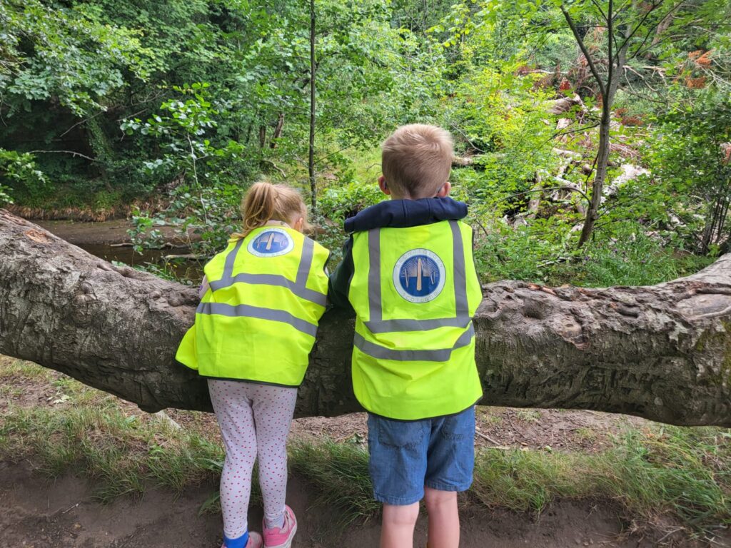Two children in hi-vis vests are leaning on a branch, with trees in the background.