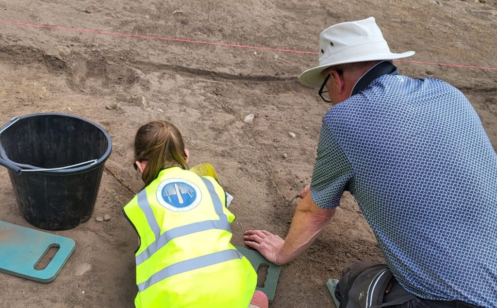 An adult and a child kneel side by side. The adult is resting their hand on the ground and the child has a black bucket next to them.