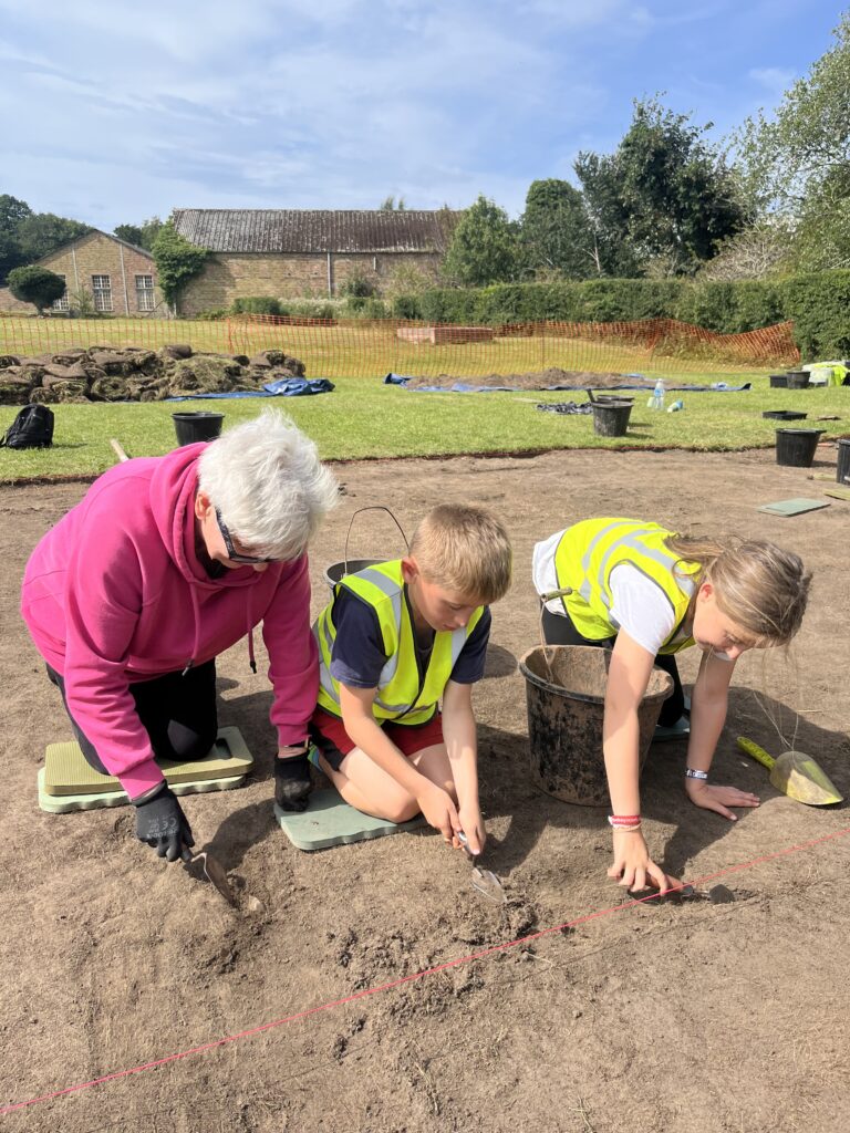 An adult in a pink hoodie and two children in hi-vis vests are kneeling on the ground in the sunshine. They are each holding a metal trowel.
