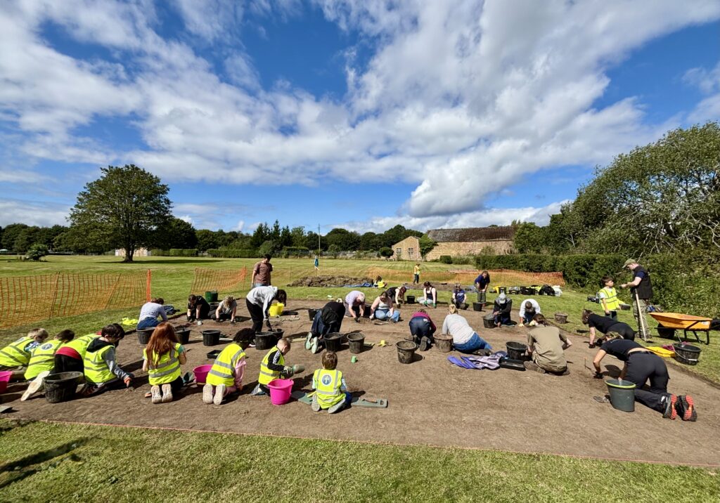 Below a bright blue cloudy sky is an open trench with brown soil. Inside the trench are lots of people kneeling, wearing hi-vis yellow vests. There are trees in the background.
