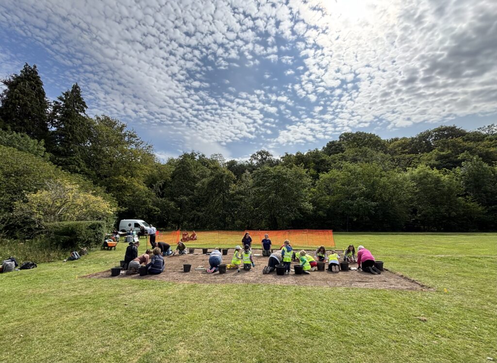 People wearing yellow hi-vis vests are kneeling in a shallow trench on the soil. Behind them is an orange fence and trees. There is grass all around the shallow trench.