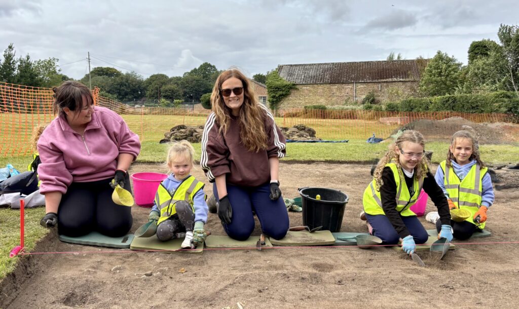 Smiling adults and children kneeling on the ground, with a pink bucket and a black bucket by them. Behind is a building.
