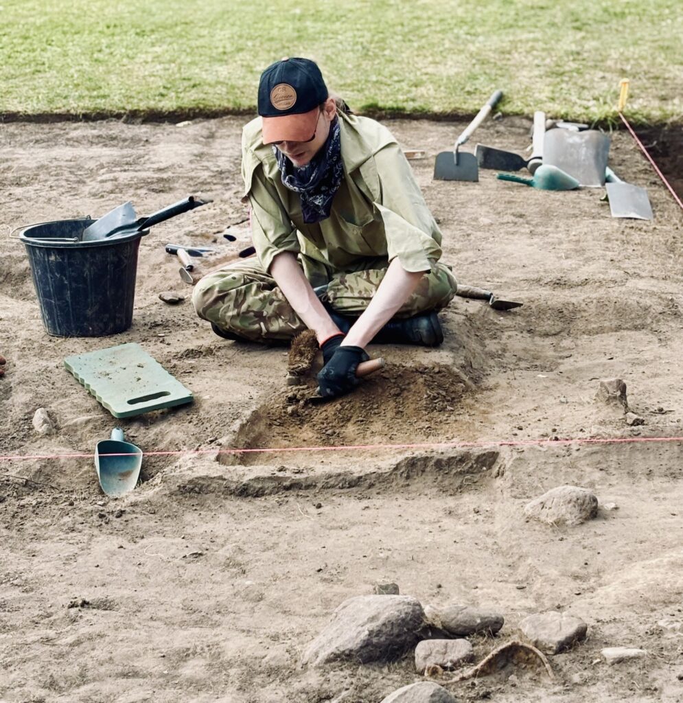A person in khaki clothes and a black and brown cap and black gloves is sitting on the ground holding a trowel. Around them are stones, a green scoop, a green kneeling pad and a black bucket with a black hand shovel.