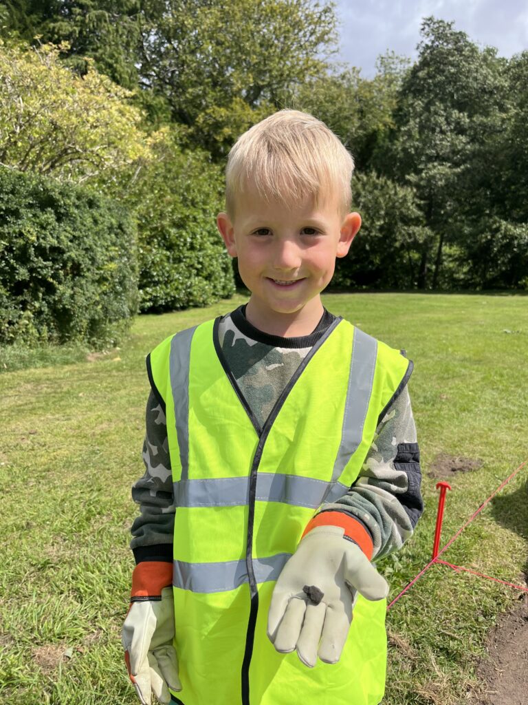 A child wearing a yellow hi-vis vest and grey gloves is smiling and holding out a small piece of coal.