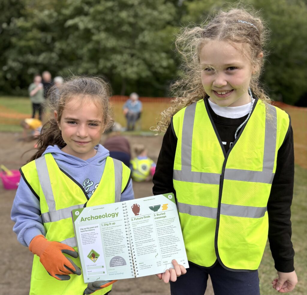 Two children are smiling and wearing yellow hi-vis vests. They are both holding up an open book with text and icons.