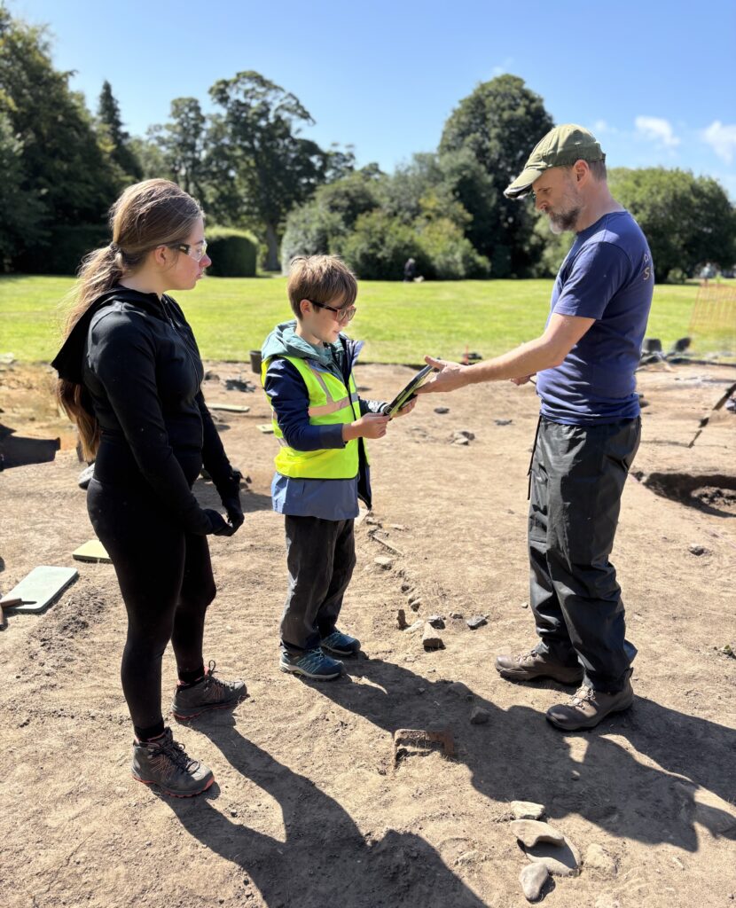 Three people are standing in the sun. The person on the right is holding out an ipad to the person in the middle. They are standing on the ground and in the background is grass, trees and a blue sky.