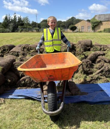 A child wearing a yellow hi-vis vest and gloves is smiling and holding an orange wheelbarrow. Behind them is a pile of turf and beyond that, grass, buildings and trees.