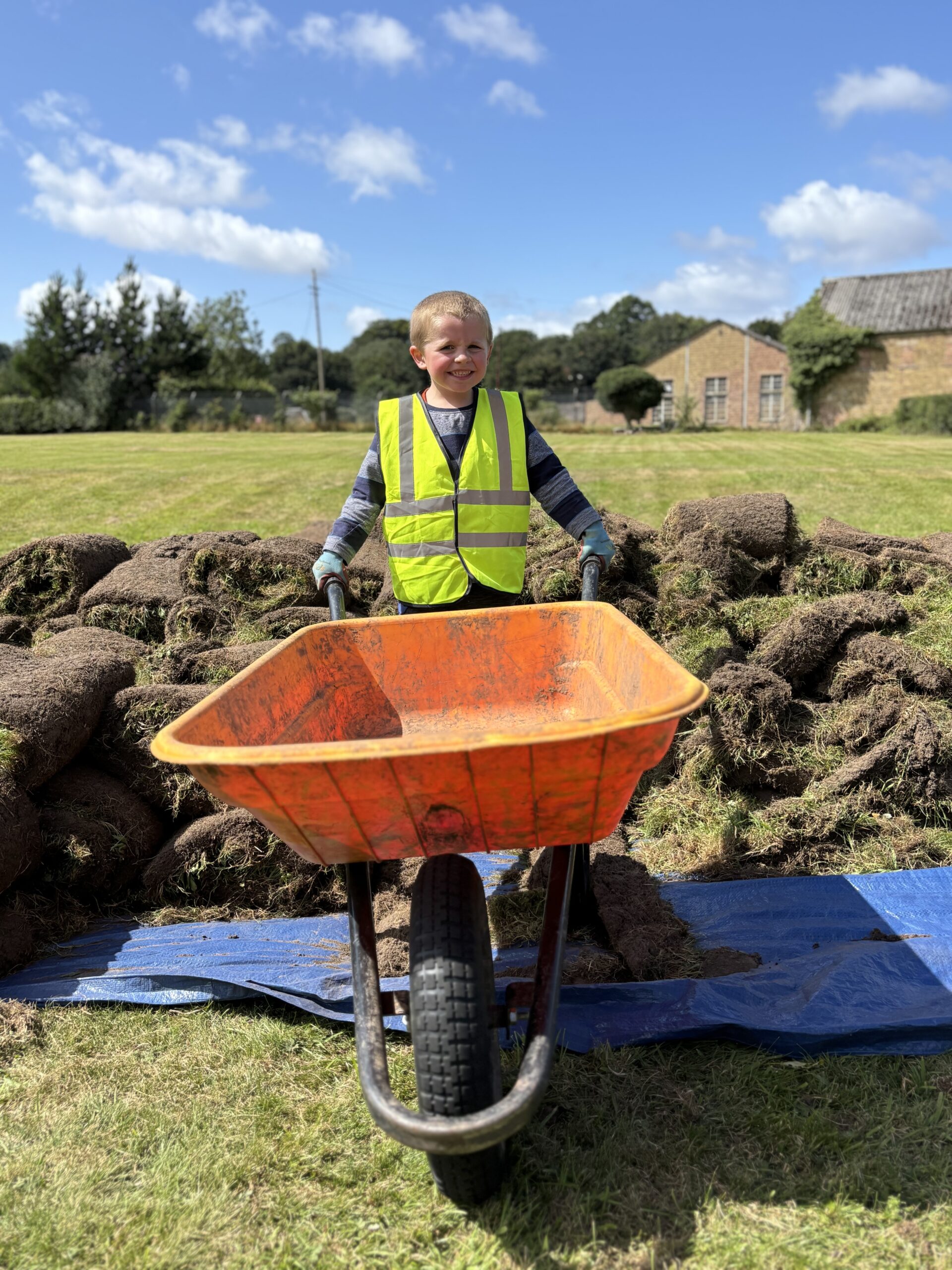 A child wearing a yellow hi-vis vest and gloves is smiling and holding an orange wheelbarrow. Behind them is a pile of turf and beyond that, grass, buildings and trees.