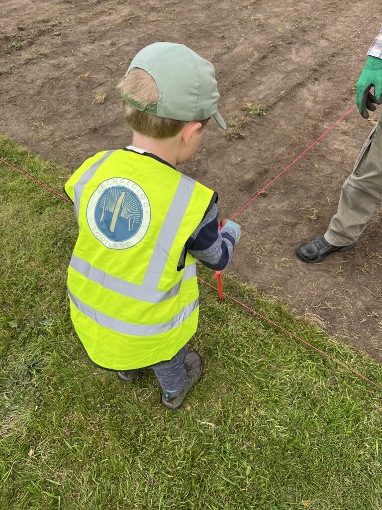 A child in a yellow hi-vis and a green cap is facing the ground. They are holding an orange string and underneath their hand is an orange peg.