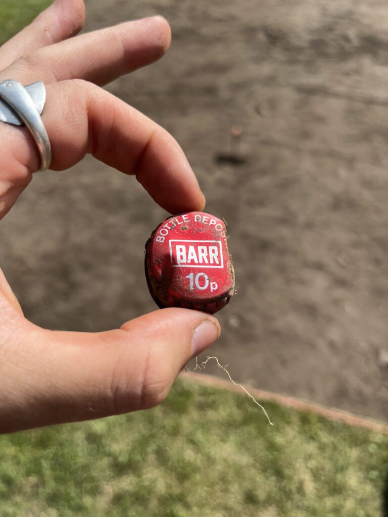A hand holds a red metal botte cap with white text.
