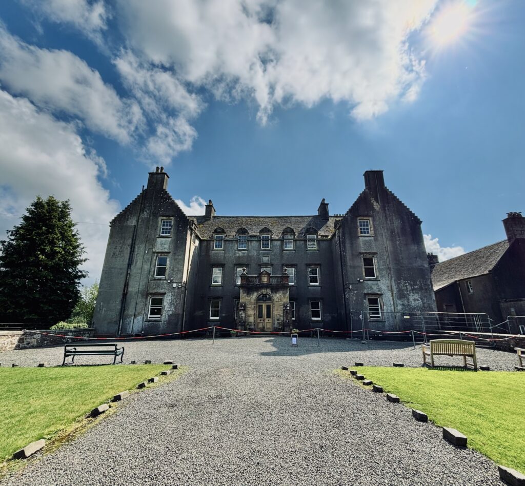 A largegrey house with lots of windows is at the end of a gravel drive, with grass either side and stone kerbs. The sun is shining and the sky is bright blue and cloudy. The focus is 0.5x