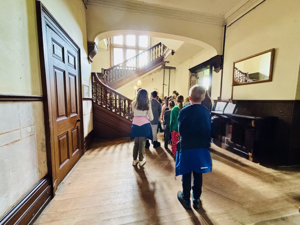 People stand looking towards a curving wooden staircase. Sun shines down from a window and there are shadows across the wooden floor.