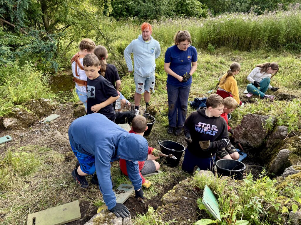 A group of people are busy digging, walking and looking at a rockery. They are wearing gloves, some are holding trowels. There are lots of plants surrounding the group and some kneeling pads scattered on the ground.