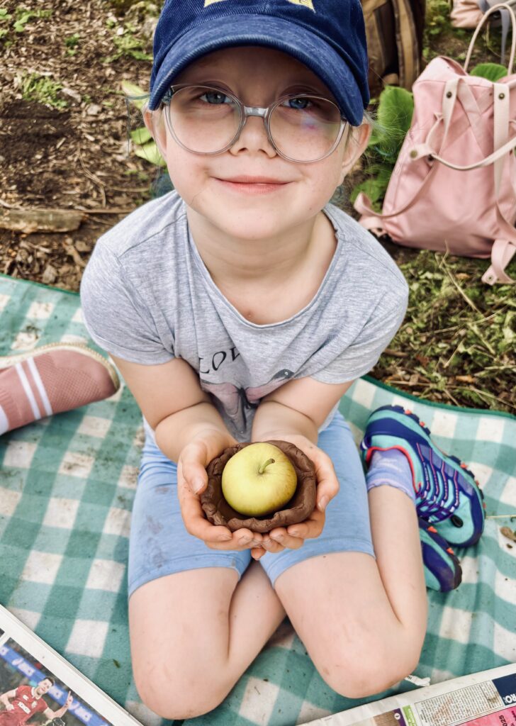 A child with glasses and a blue cap is holding a pot in both hands, with an apple inside. They are sitting on a blue and white chequered blanket