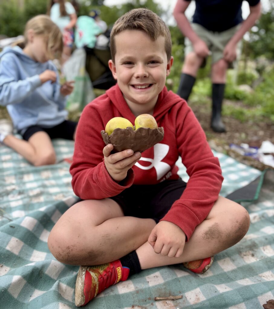 A child in a red hoodie is smiling and holding a pot in one hand, which has two yellow apples inside. They are sitting on a blue and white chequered blanket and there are out of focus people in the background.