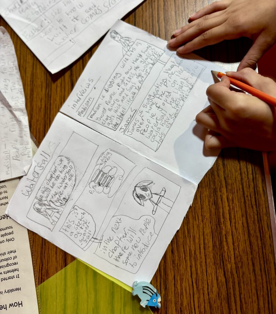 A hand drawn double page comic is open on a brown wood pattern table. In the corner, someone is holding the page steady with one hand while they write with their other hand using an orange pencil.