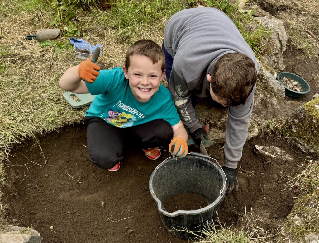 One person in a grey hoody on the right is kneeling and using a scoop next to a bucket. On the left, a boy in a turquoise tshirt and orange gloves is smiling and showing a thumbs-up. Behind them is grass and soil.