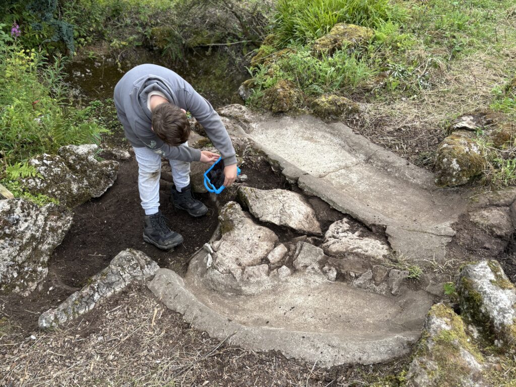 A boy is standing bent over holding an ipad in both hands. Around him is a concrete feature that is gently curved and sloping. There are rocks and plants.