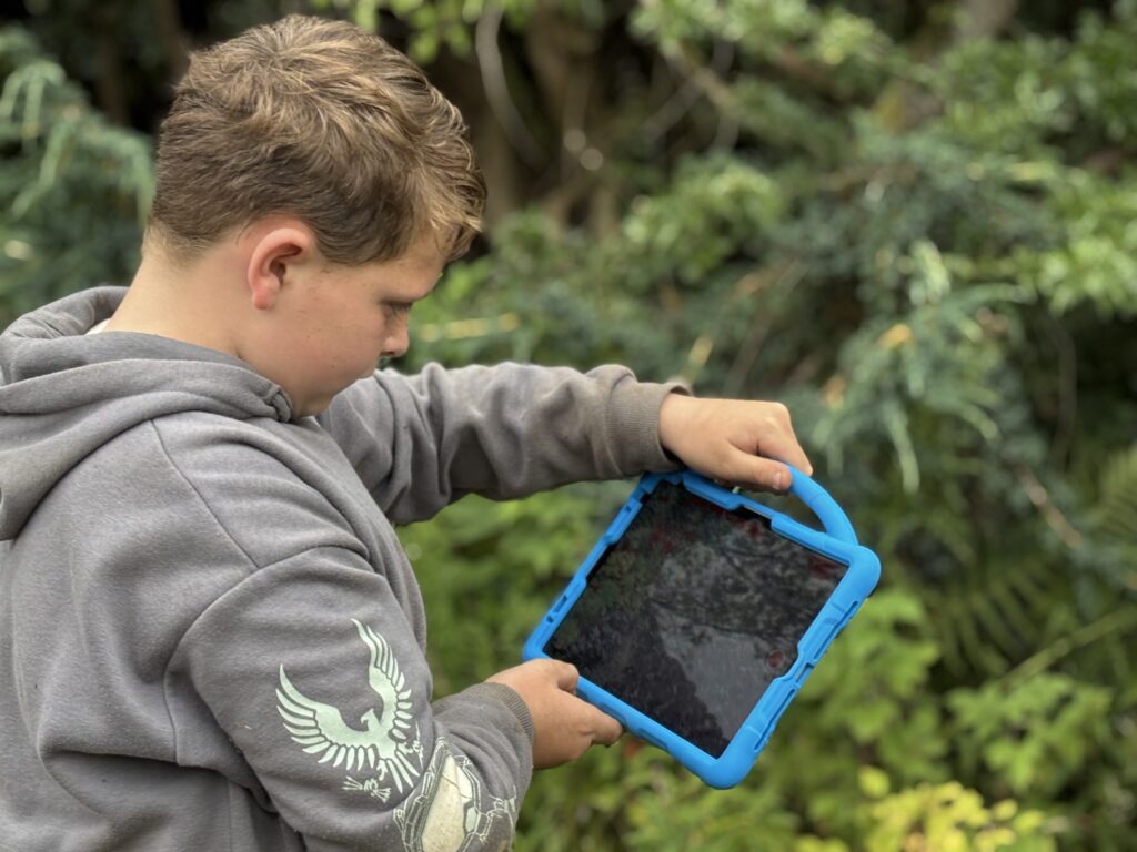 A boy in a grey hoody is holding an ipad with a blue case and handle. He is holding it with two hands. The background is out of focus plants.