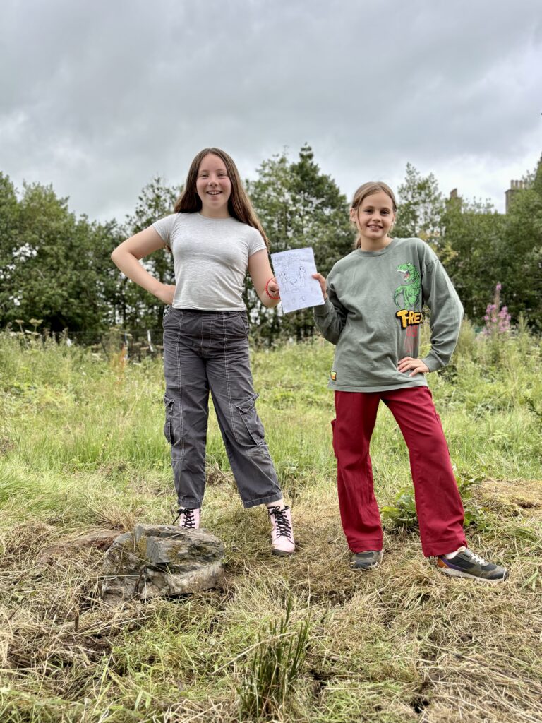 Two smiling girls are standing on grass with trees in the background. They have one hand on their hip, and with their other hand they are both holding onto a comic.