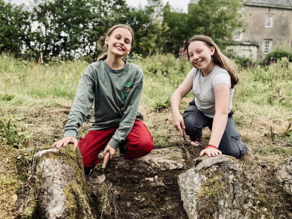 Two smiling people are kneeling on mossy rocks. They are each holding a trowel. In the background out of focus are trees and a grey house with windows.