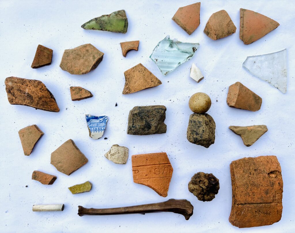 On top of a white background are lots of pieces of broken objects. Some are reddish orange, some are clear glass and some are black and brown. They are different shapes and sizes, including one round marble ad one animal bone.