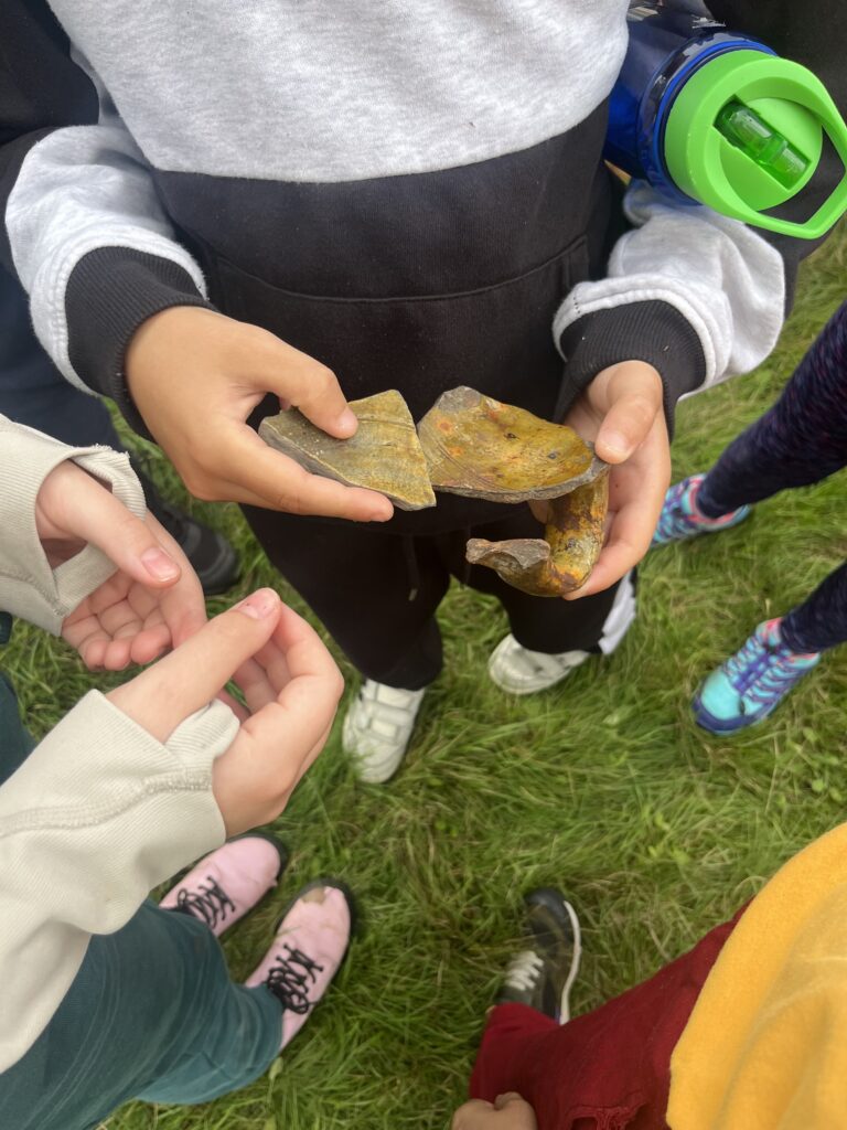 In the centre, someone is holding two pieces of green pottery in their hands. One has a handle. There are other hands reaching out to hold it, and more people are standing in the background with their shoes in the photo.