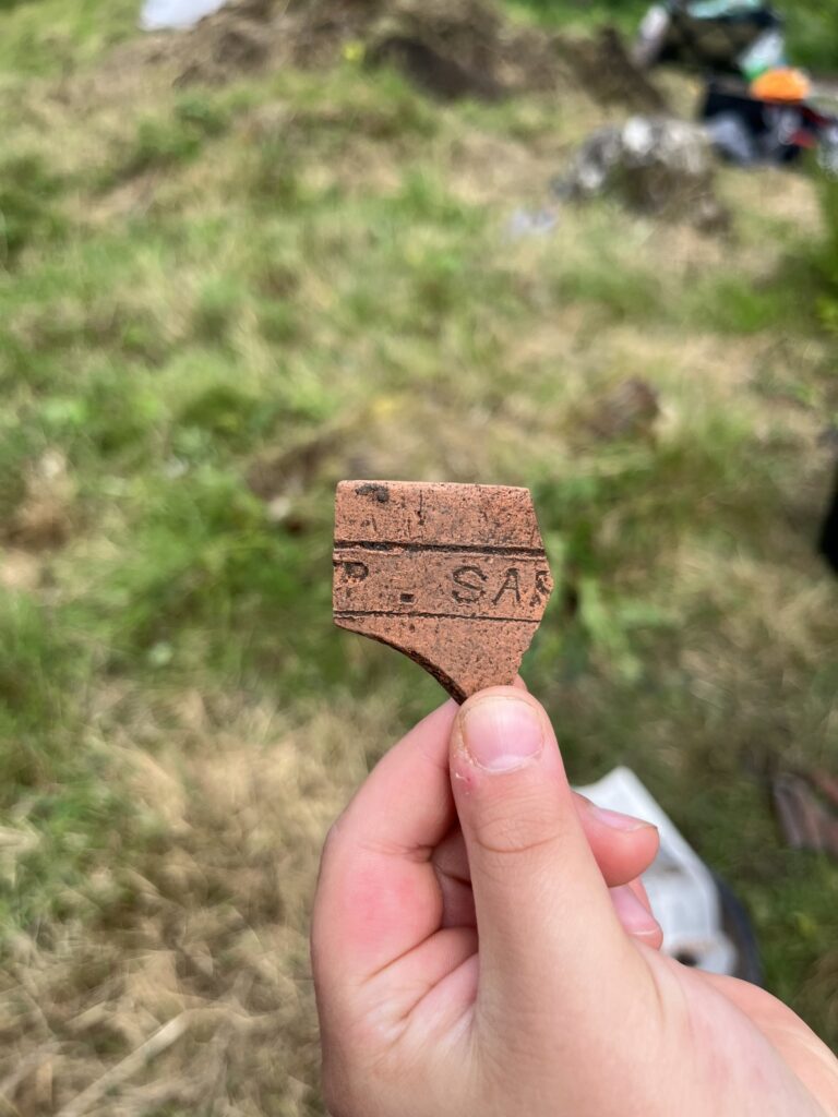 a hand is holding up a dirty piece of reddish orange pottery using their finger and thumb. The pottery has the letters 'SA'. The background is out of focus grass.