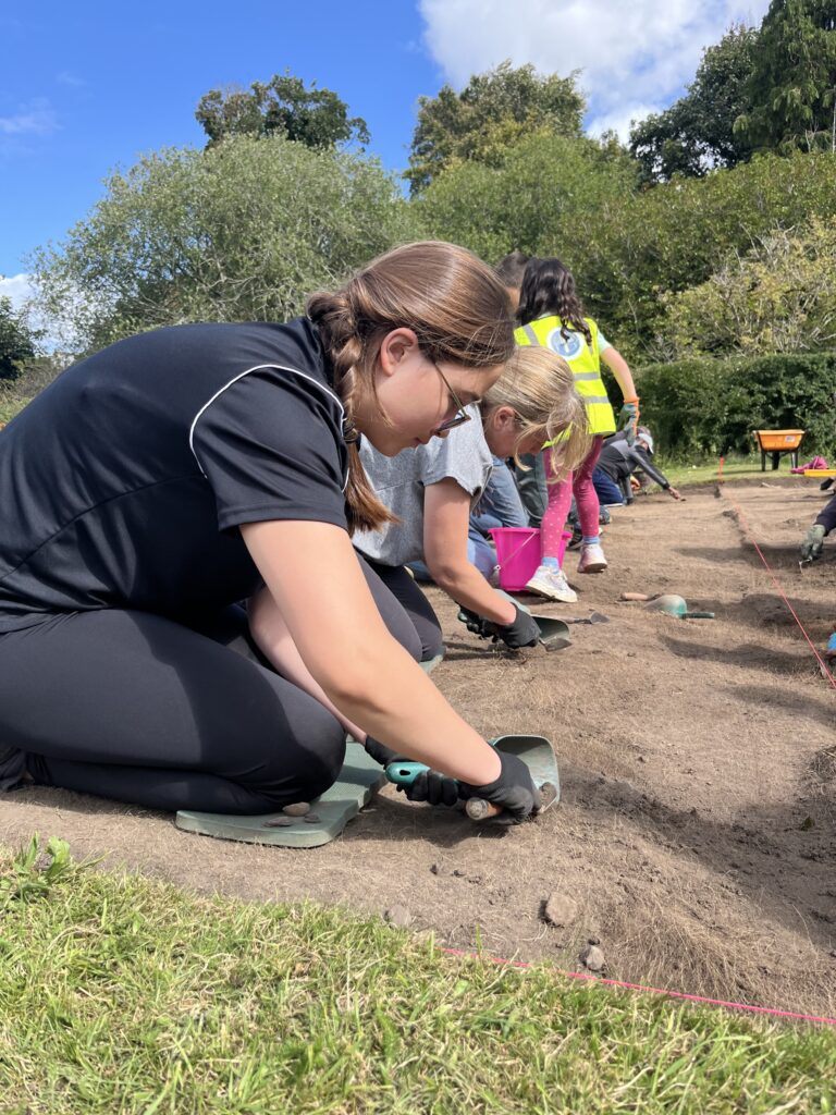 A row of people kneeling on the ground. A person wearing a black tshirt and gloves is holding a trowel.