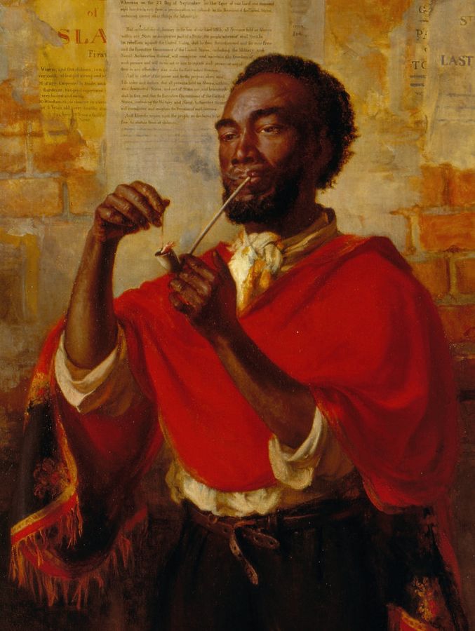A man is holding a long pipe in one hand and lighting it using a match. He is wearing a red loose garment and a white shirt underneath with the sleeves rolled up. Behind him is a brick wall with torn posters.