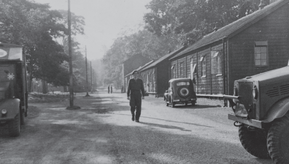 A smiling man is walking along a driveway, with huts on one side and vehicles on both sides. There are trees on each side, and telegraph poles on the left.