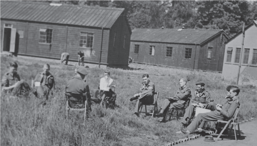 A black and white photo oh wooden huts in the background, with a group of people wearing uniform sitting around one person in a hat.