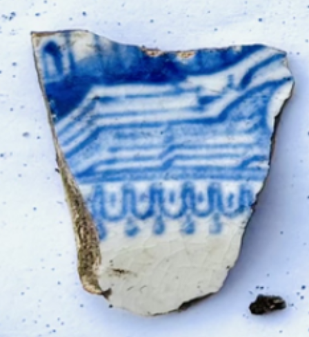A slightly blurry photo of a white piece of pottery with dirt on one side. It is covered in a blue pattern with straight and curved lines and shading. The pattern is not clear.