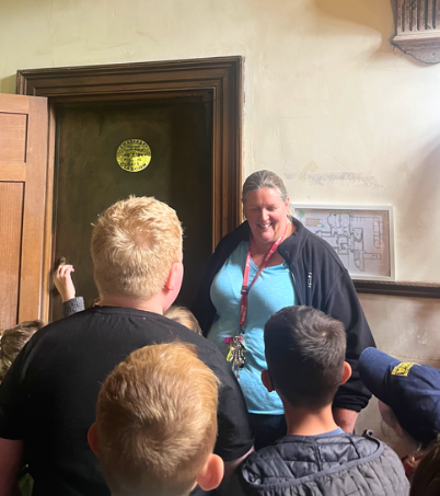 A smiling woman is looking at a group of children. She is wearing a blue tshirt and a black fleece. Behind her is a big brown door with a gold sign on it.