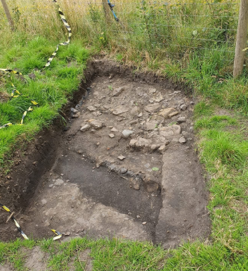 An open trench is surrounded by grass, with some hazard tape to the left. Inside the trench there is a dark gully, and on one side there are lots of stones.