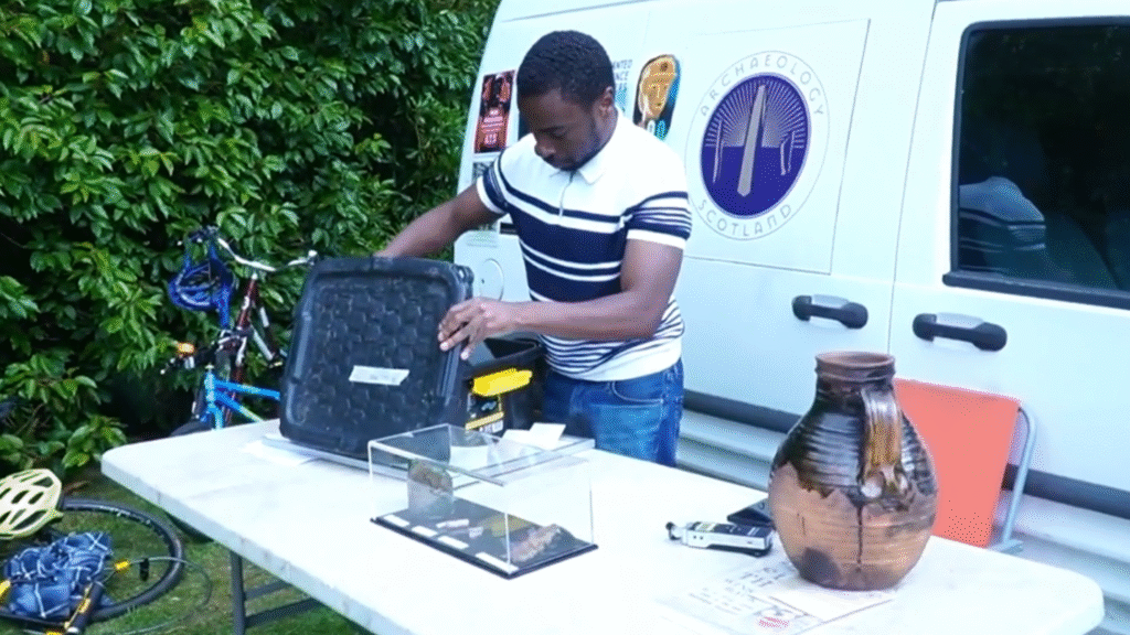 A person is looking inside a box on a table that has a jug and other small objects. Behind them is a white van with a logo and a bush.