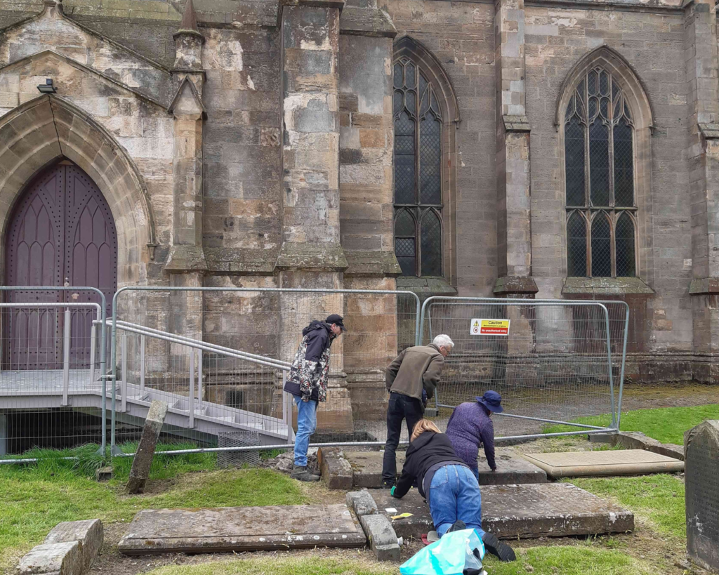 Four people at a graveyard in front of an abbey. Three of the people look and point at a gravestone; the fourth person kneels down and works on a gravestone.
